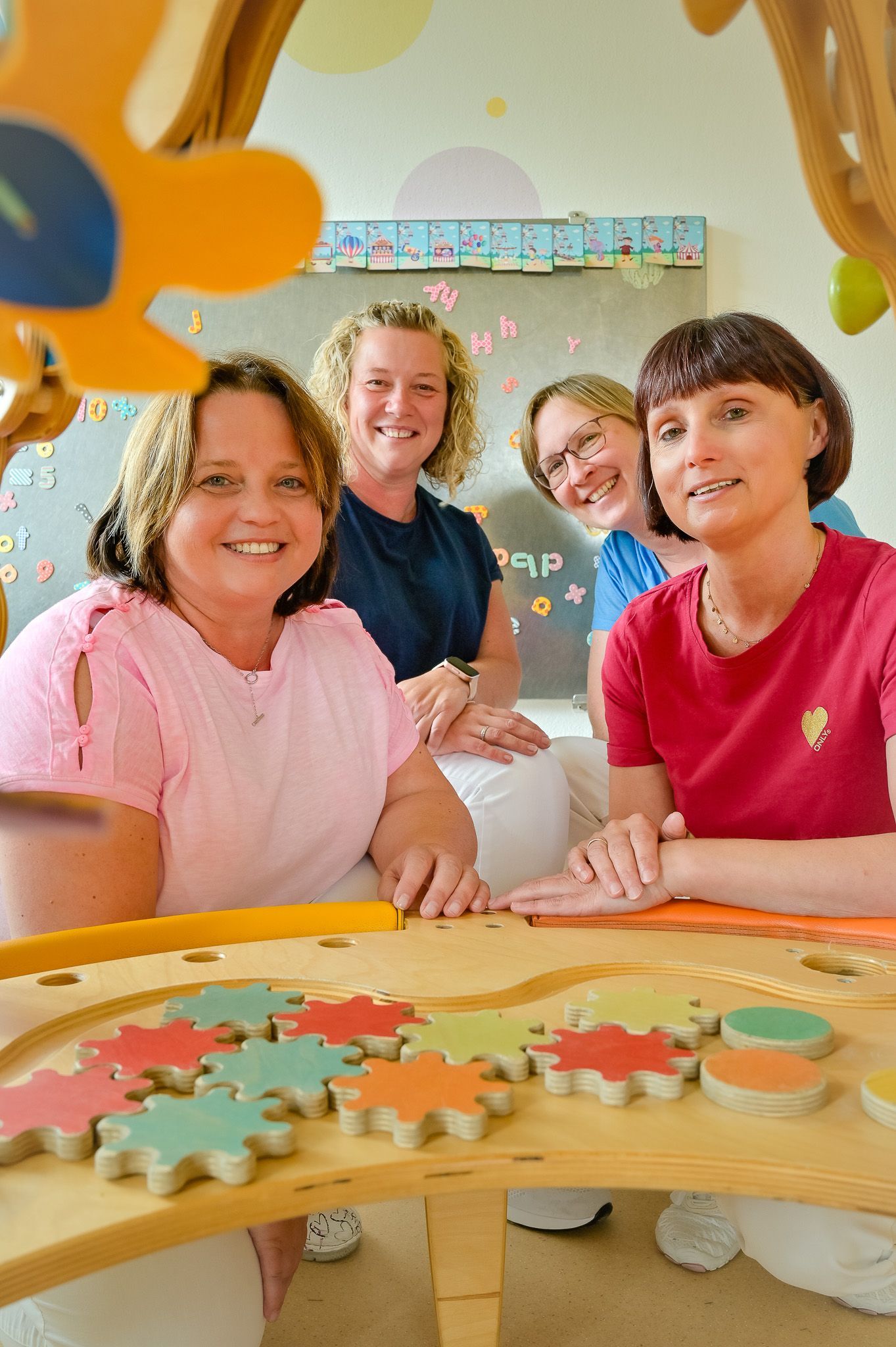 Vier Frauen lächeln an einem Tisch mit bunten Holzspielzeugen in einem Klassenzimmer.