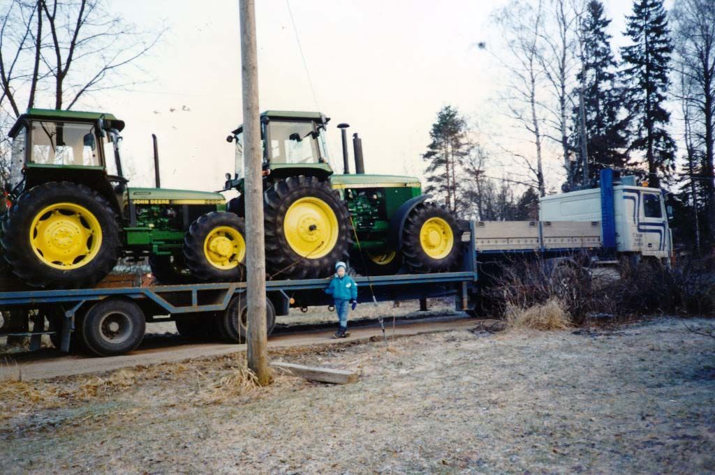 Kaksi John Deere -traktoria perävaunussa, ihmisen ja kuorma-auton vieressä. 