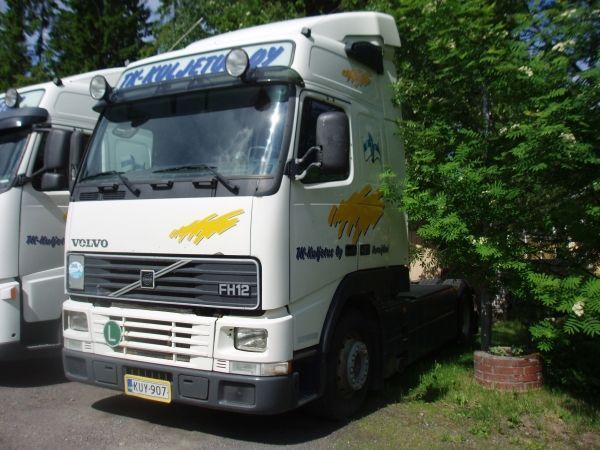 Valkoinen Volvo FH12 -kuorma-auto pysäköitynä vihreän pensaan viereen; keltainen logo kyljessä; sininen taivas.
