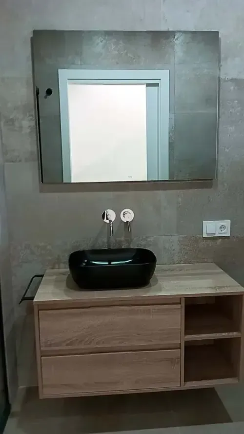 Baño con lavabo negro sobre tocador de madera, espejo grande y azulejos de pared color gris claro.