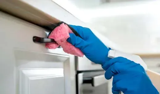 Persona con guantes azules, limpiando la manija de la puerta de un armario blanco con una esponja rosa.