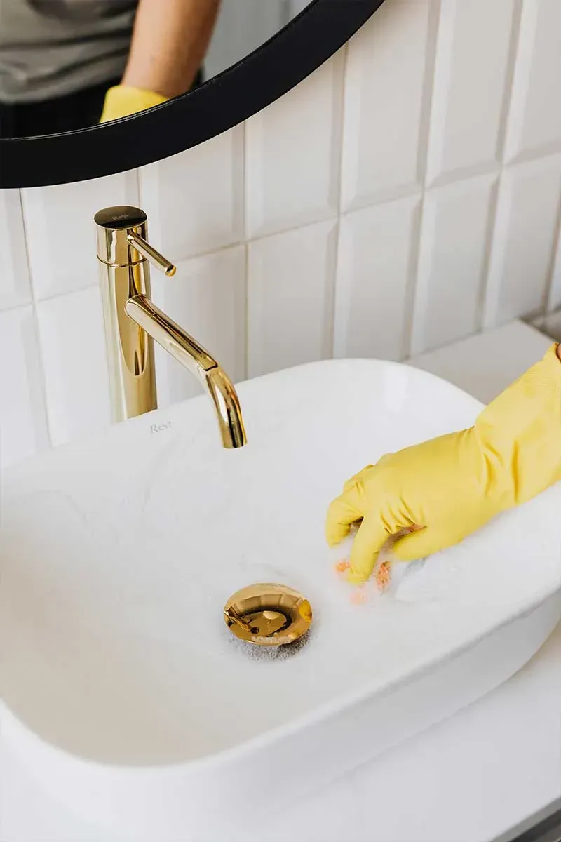 Persona con guantes amarillos limpiando un lavabo de baño blanco con un grifo dorado.