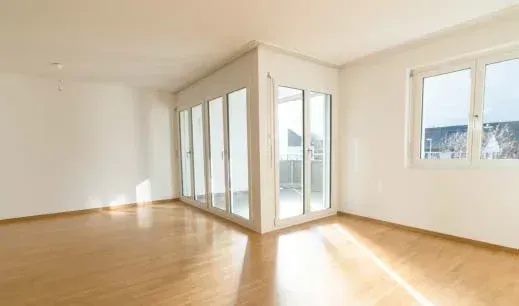 Habitación vacía y luminosa con suelos de madera y grandes ventanales.