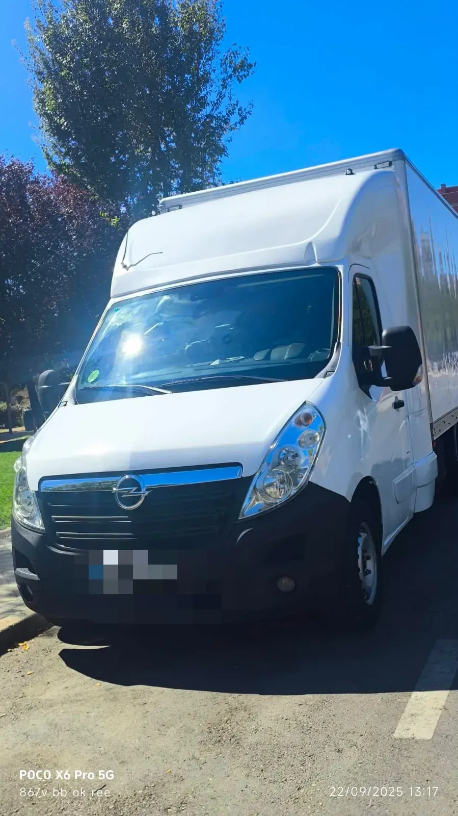 Furgoneta de carga blanca estacionada en una calle soleada, árboles y edificio al fondo.