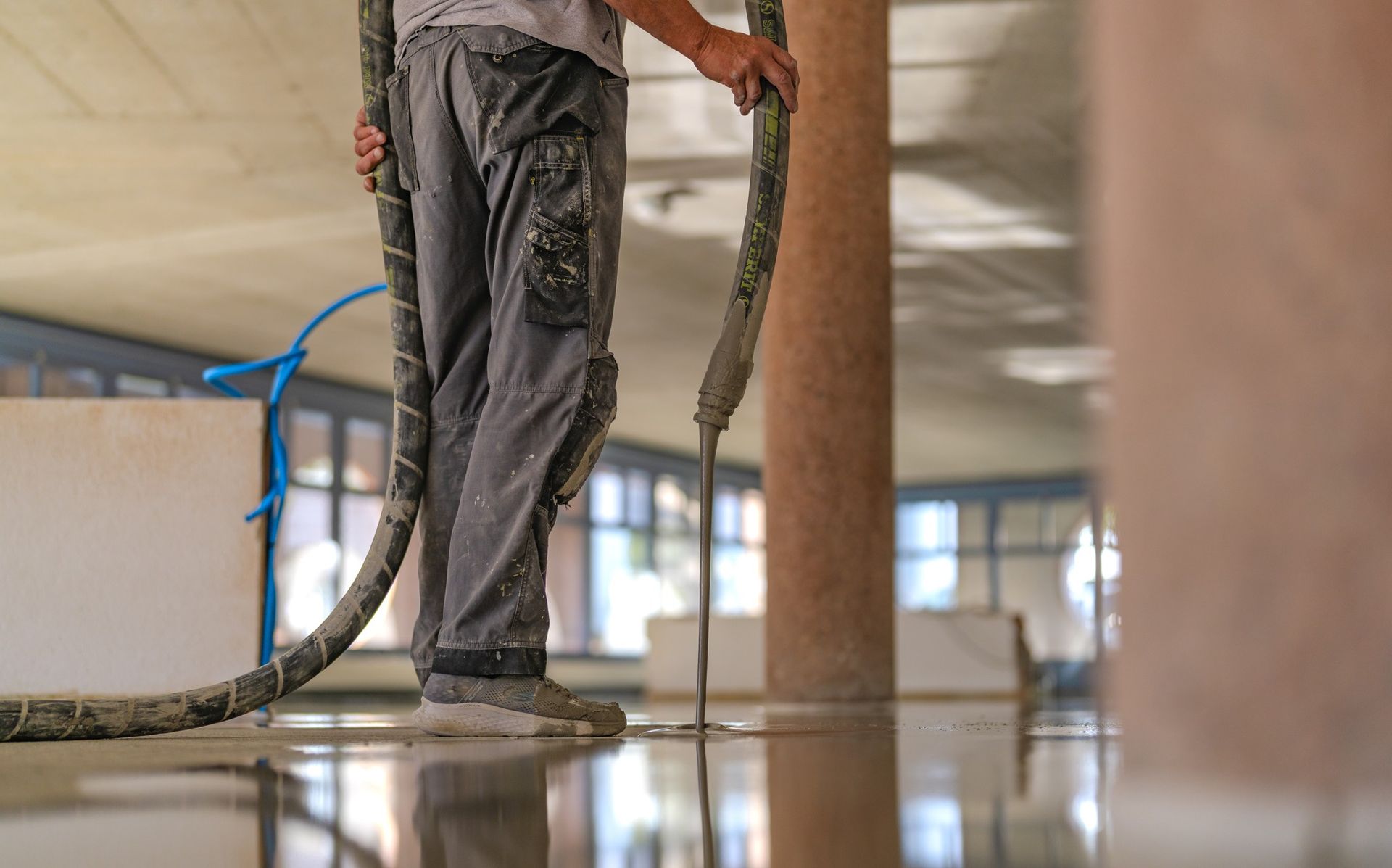 À l'intérieur d'un bâtiment, un ouvrier vêtu d'un pantalon de travail gris verse un enduit autonivelant sur une surface en béton à l'aide d'un tuyau.