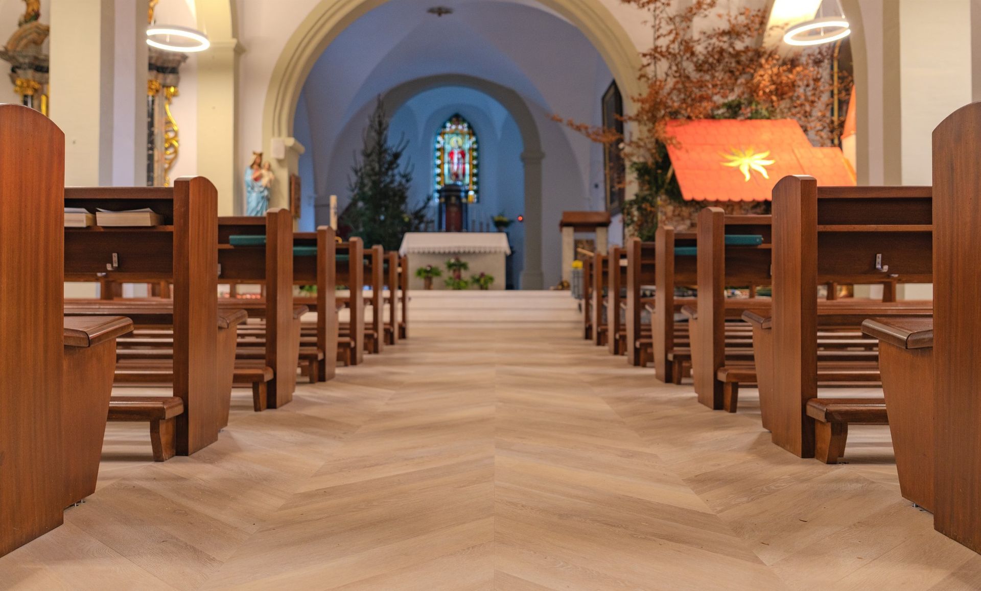 Vue intérieure de la nef d'une église avec des rangées de bancs en bois faisant face à un autel, un sapin de Noël et un vitrail.