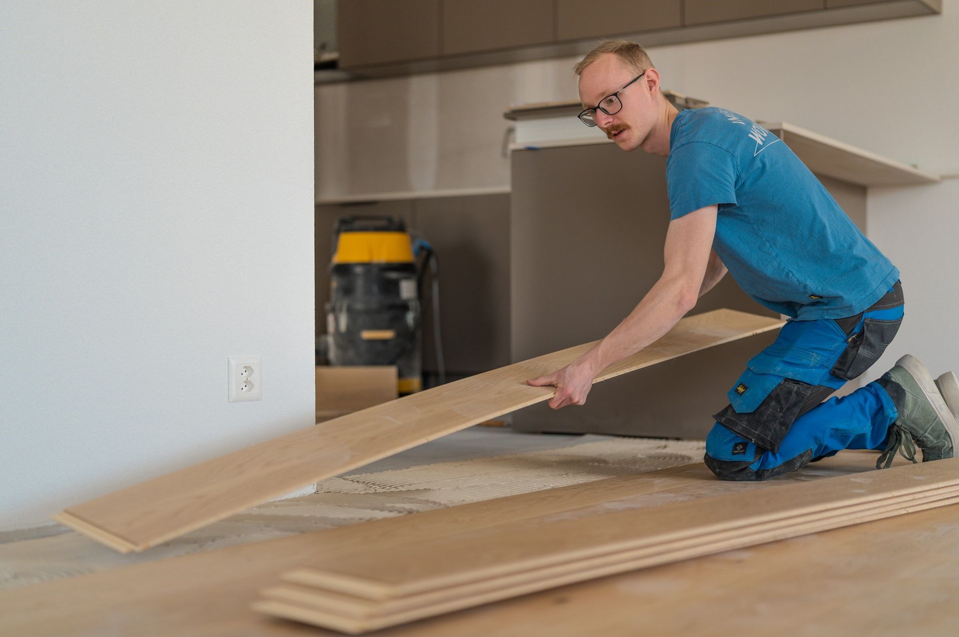 Une personne vêtue d'un t-shirt bleu et d'un pantalon de travail est agenouillée sur le sol, en train d'installer des lames de parquet clair.