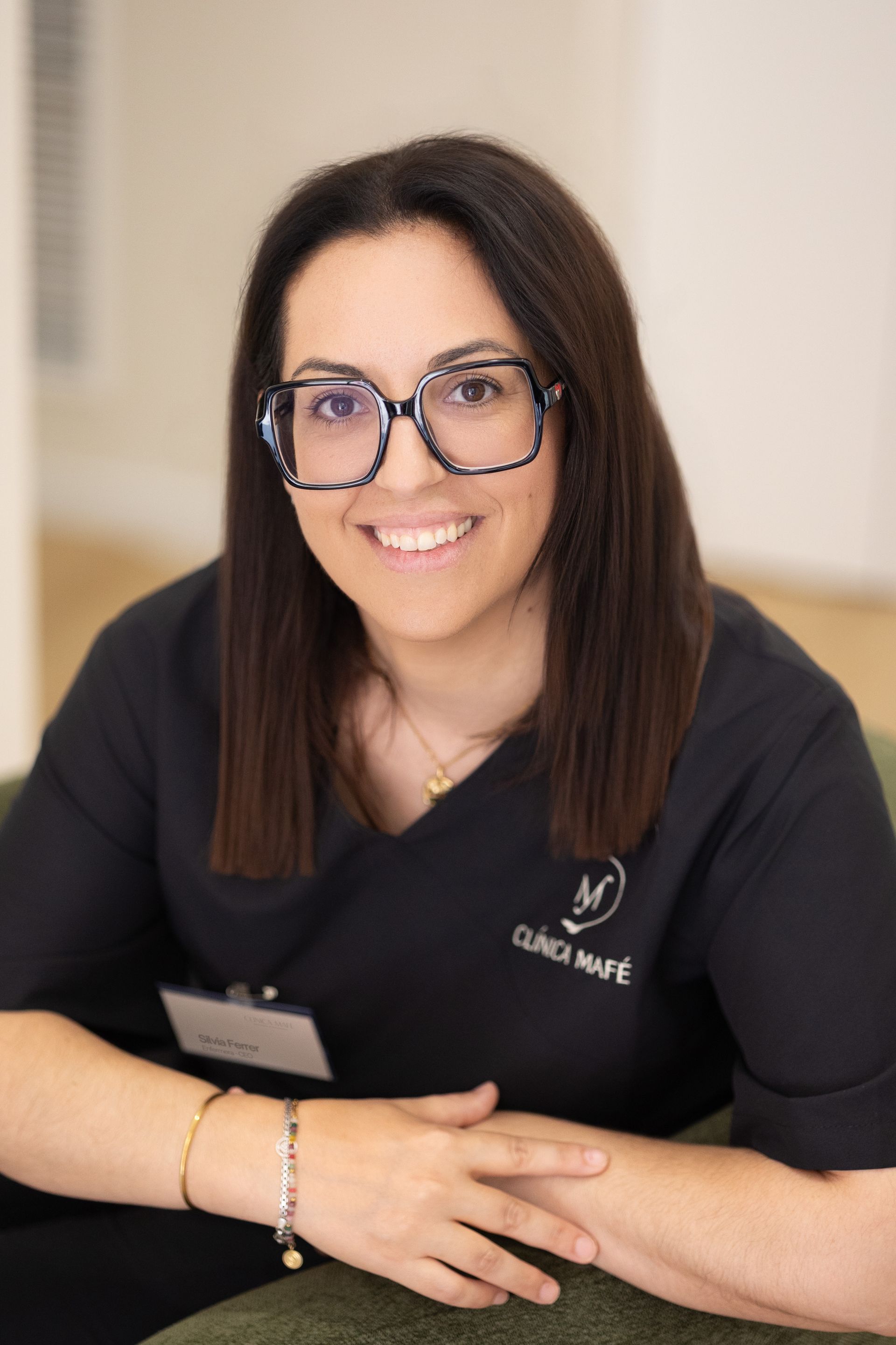 Persona vestida de negro y con gafas, sentada sobre un fondo blanco, sonriendo a la cámara.
