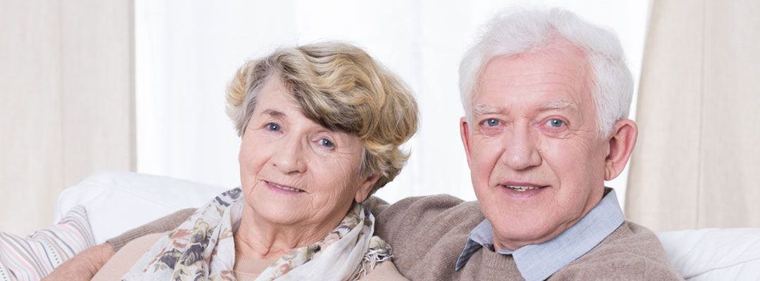 Zwei Personen sitzen eng beieinander auf einem Sofa und lächeln in die Kamera in einer hellen Innenraumumgebung.