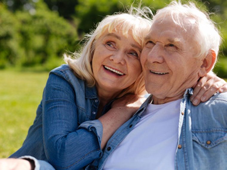 Zwei Menschen lehnen sich mit glücklichen Gesichtsausdrücken aneinander, draußen in einer grünen, sonnigen Umgebung.