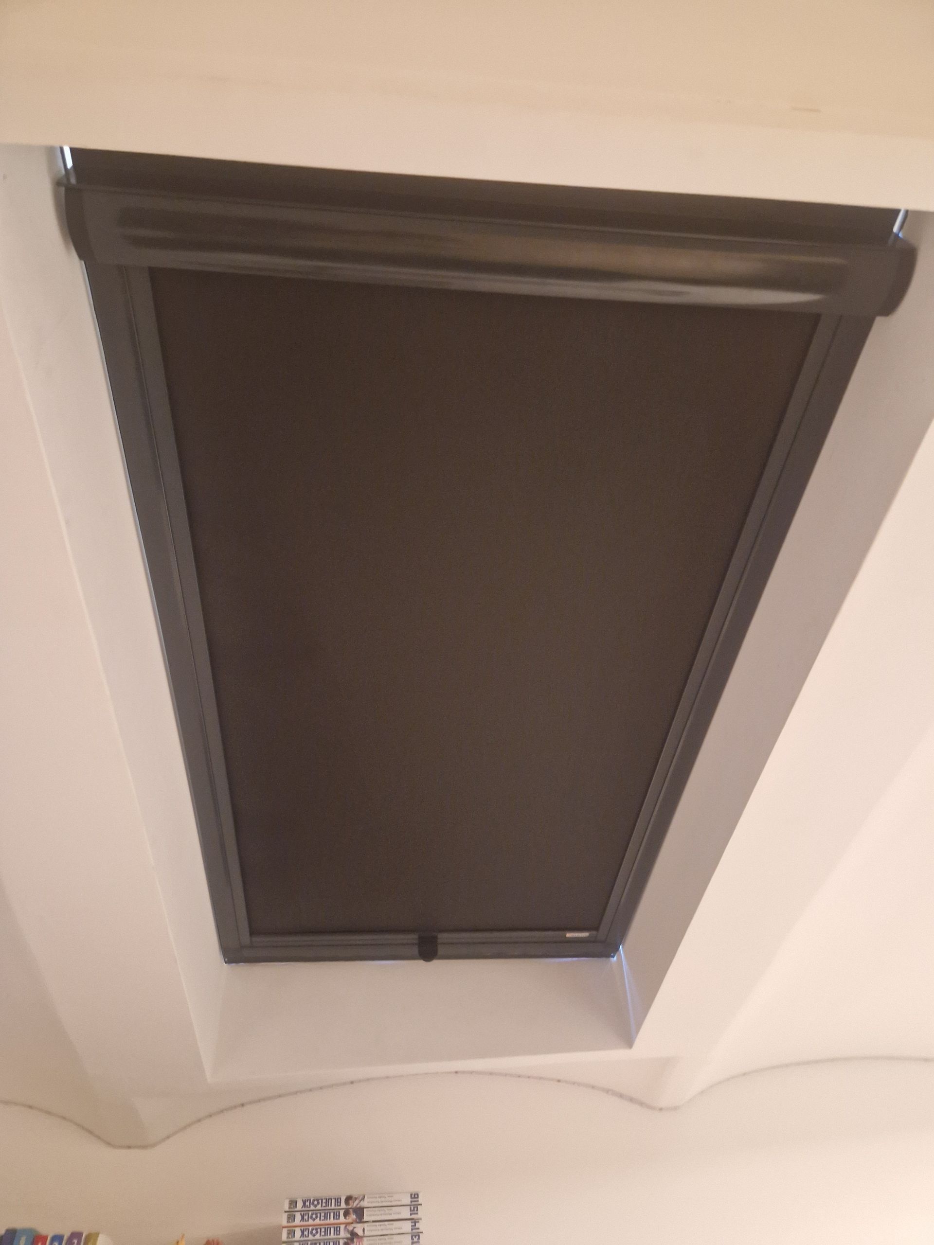 Un store occultant fermé et de couleur sombre, installé sur une fenêtre de toit encastrée dans un plafond blanc.