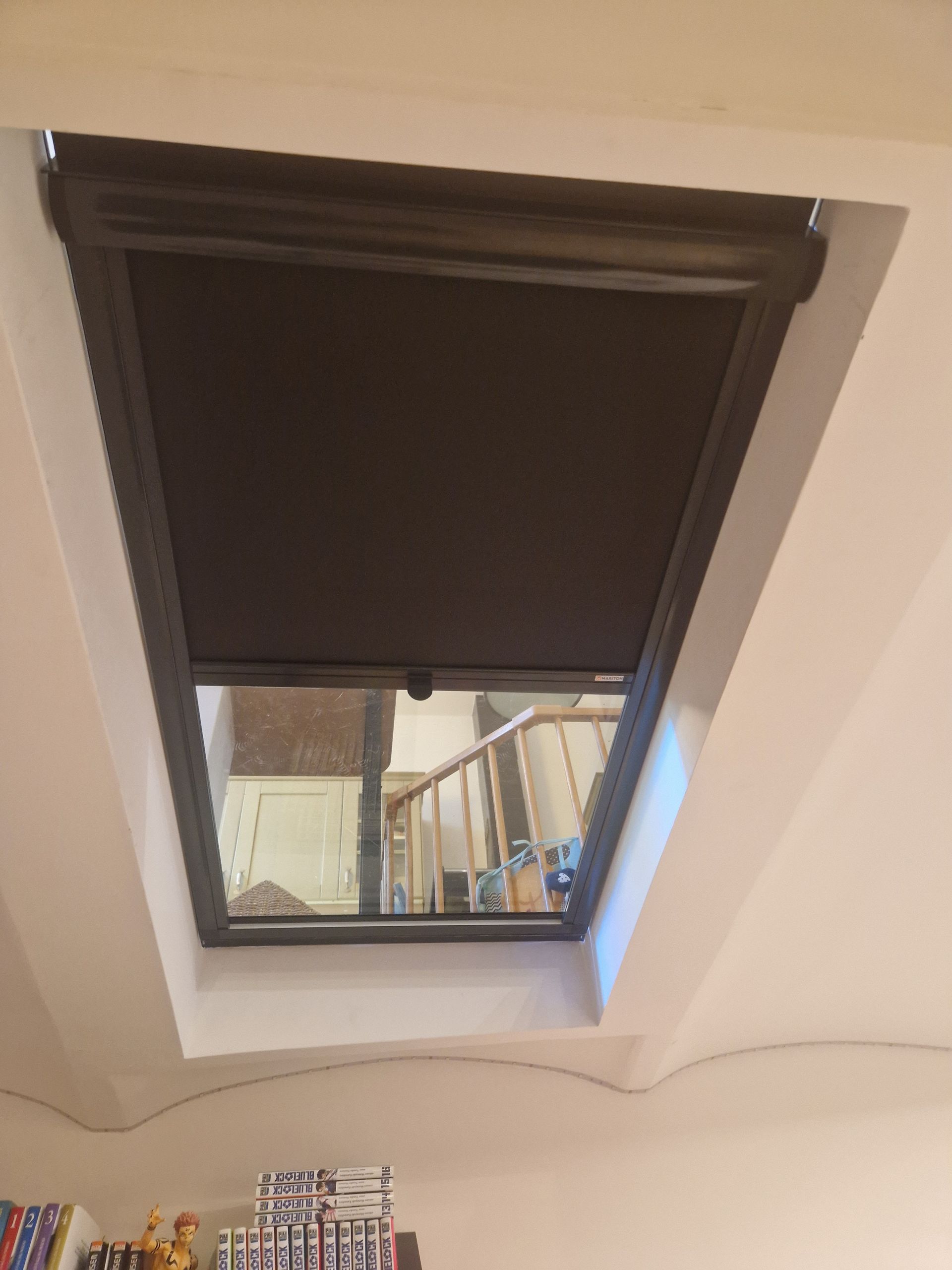 Un store occultant de couleur sombre recouvre partiellement une fenêtre de toit encastrée dans un plafond blanc, laissant apparaître un escalier en contrebas.