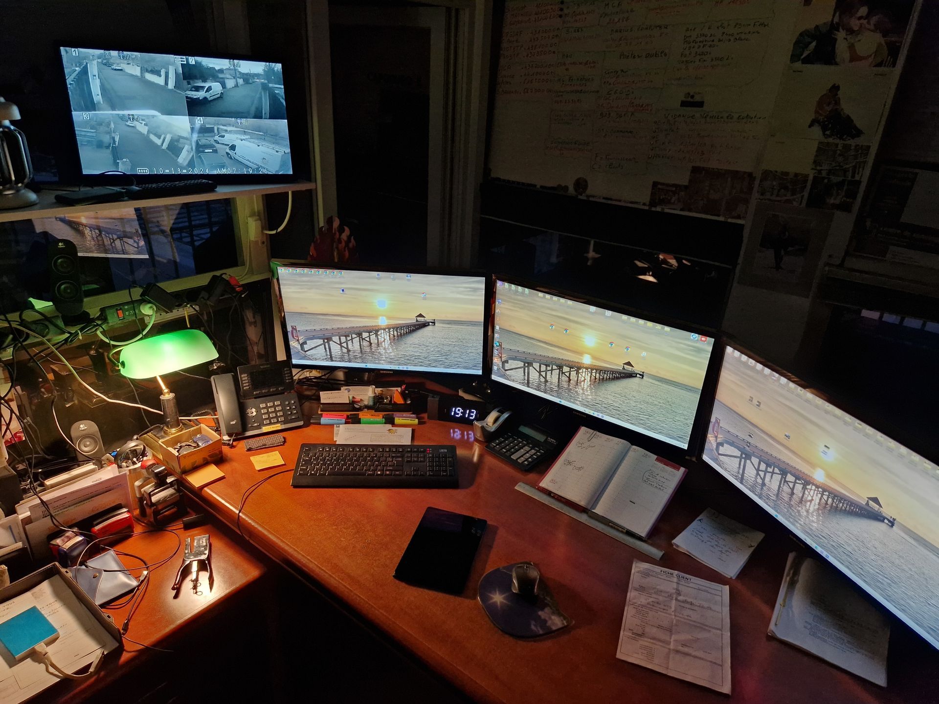 Un espace de travail faiblement éclairé avec trois écrans, un clavier, une lampe de bureau et divers papiers éparpillés sur un bureau en bois.