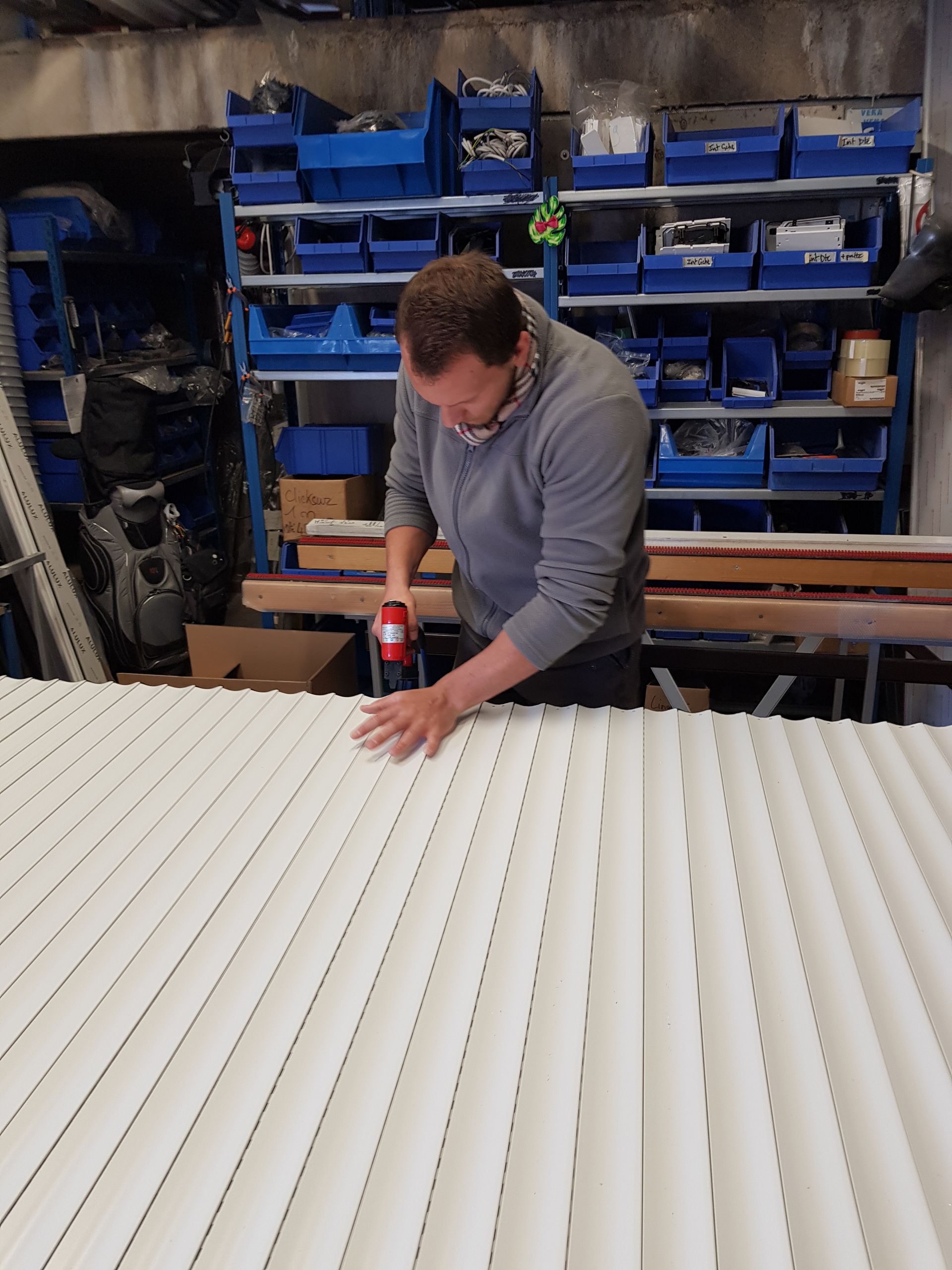 Un ouvrier en pull gris utilise un outil électrique sur un grand panneau industriel blanc côtelé dans un entrepôt.