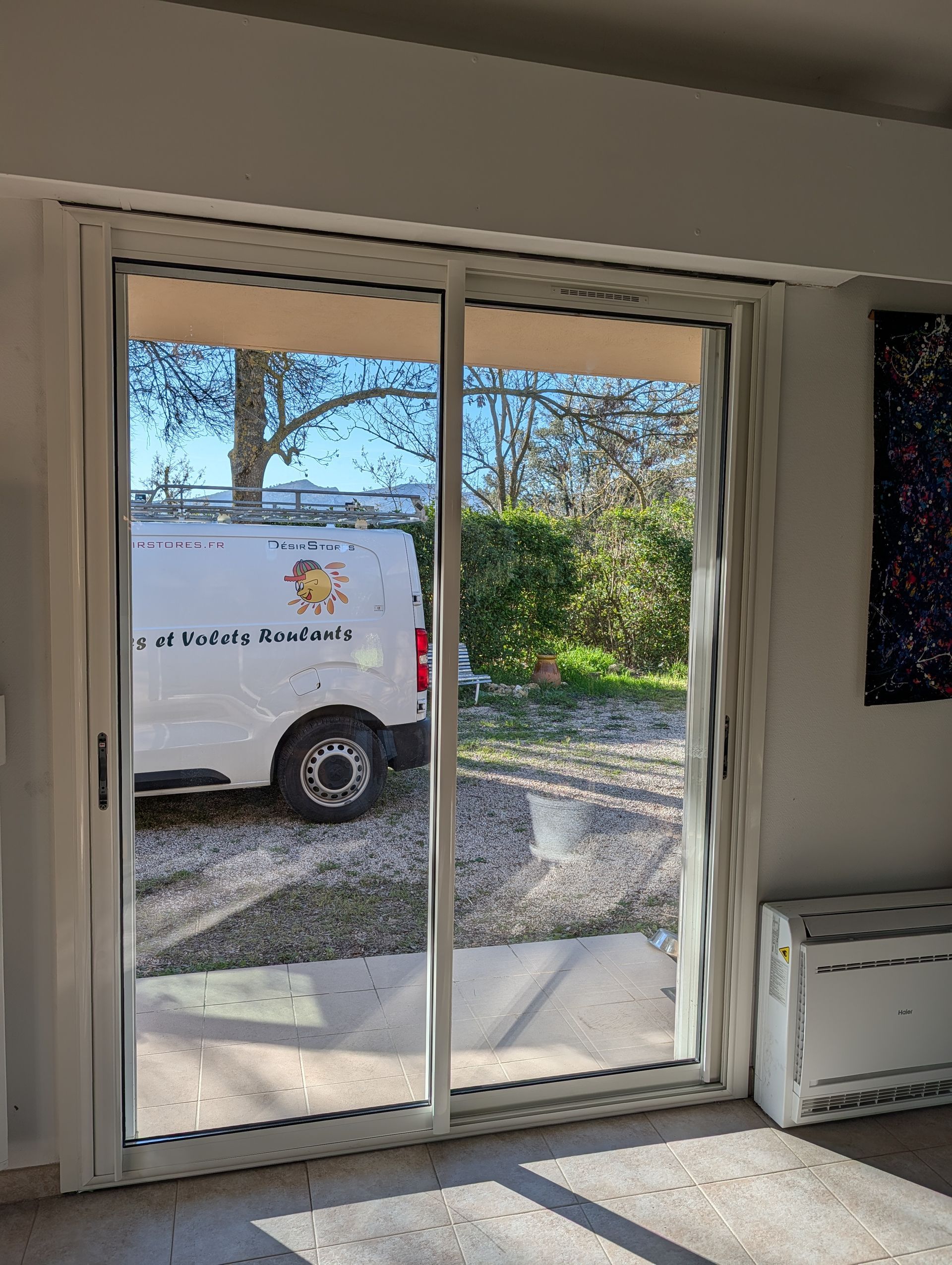 Une porte coulissante en verre blanc donne sur une allée de gravier où une camionnette blanche est garée devant des arbres.