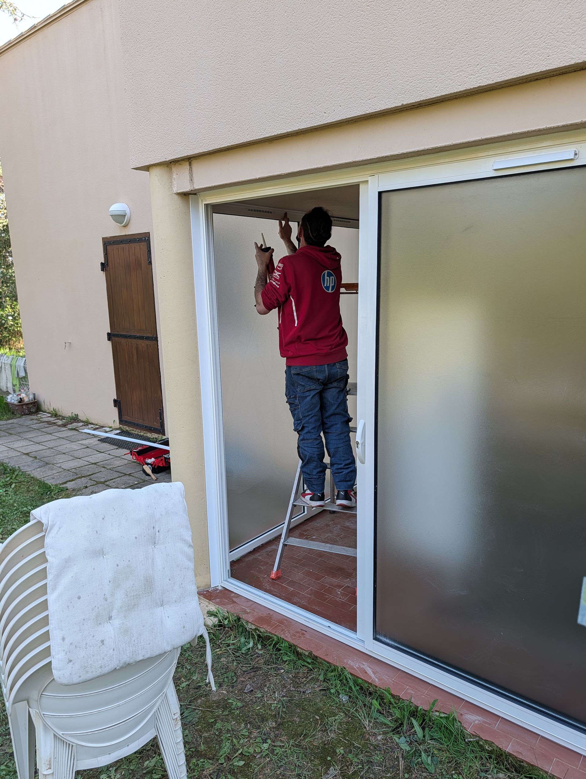 Une personne vêtue d'un sweat à capuche rouge se tient sur un escabeau, en train de peindre le cadre d'une porte-fenêtre coulissante sur la façade d'un bâtiment.