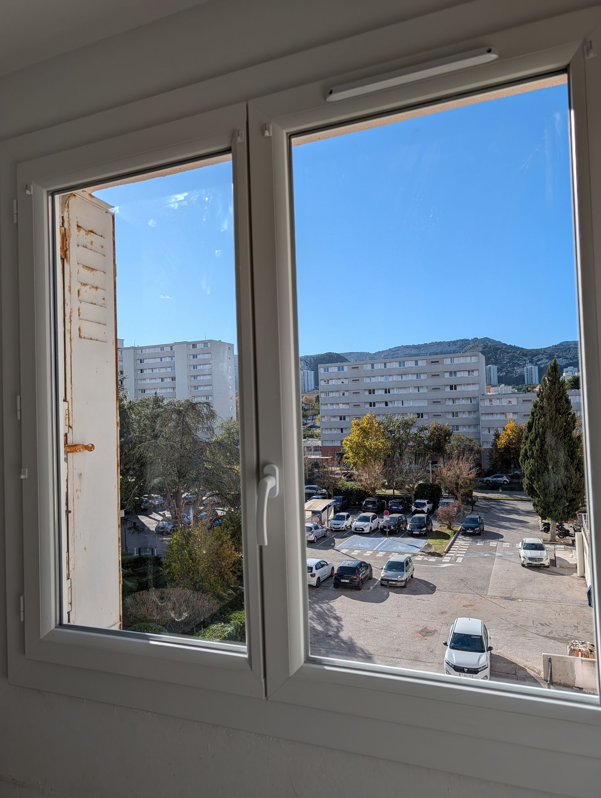 Vue à travers une fenêtre à cadre blanc donnant sur un parking résidentiel arboré et bordé d'immeubles d'appartements.