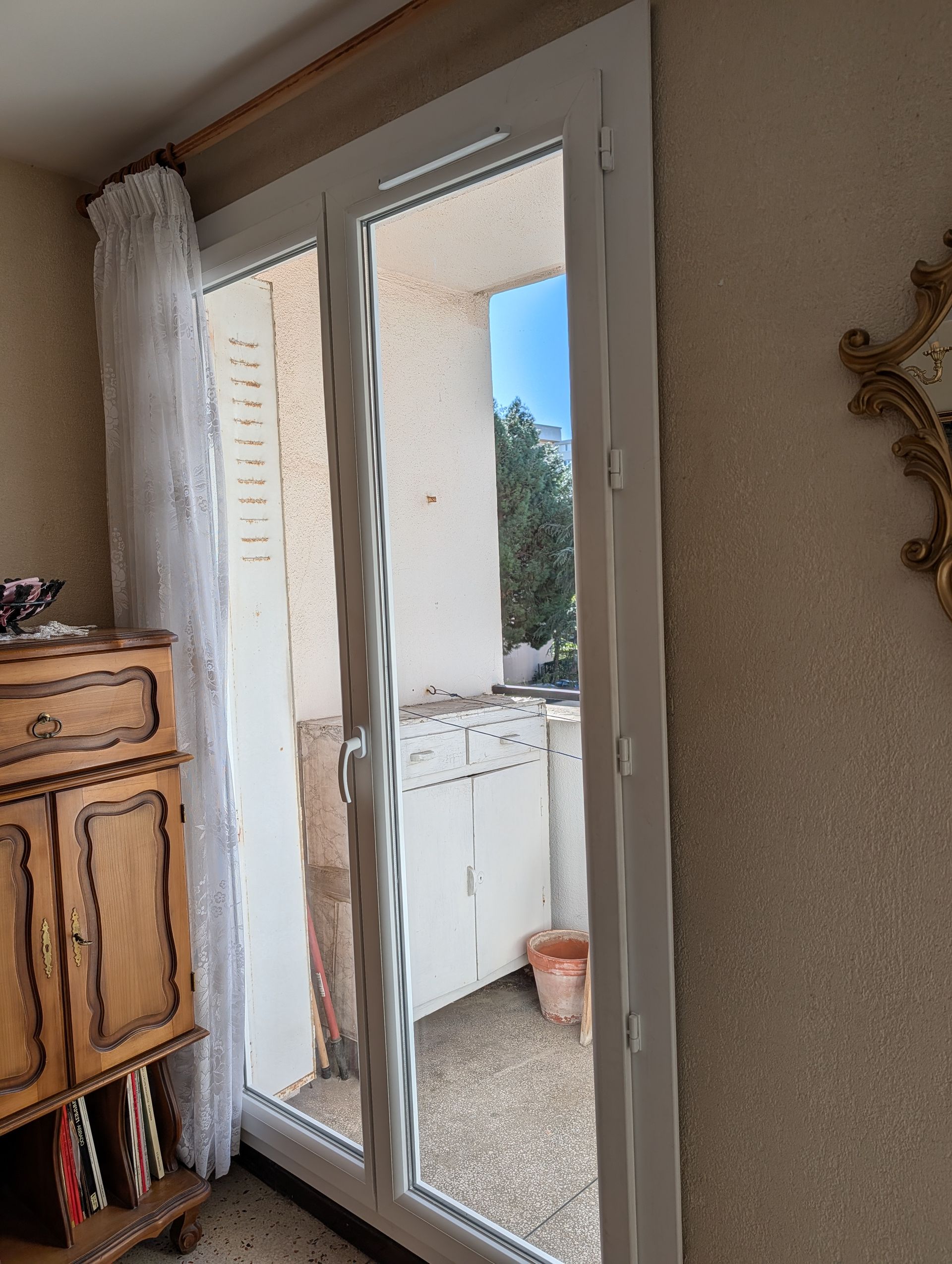 Une porte vitrée à cadre blanc donnant sur un balcon avec une petite table, un pot de fleurs et une vue sur l'extérieur.