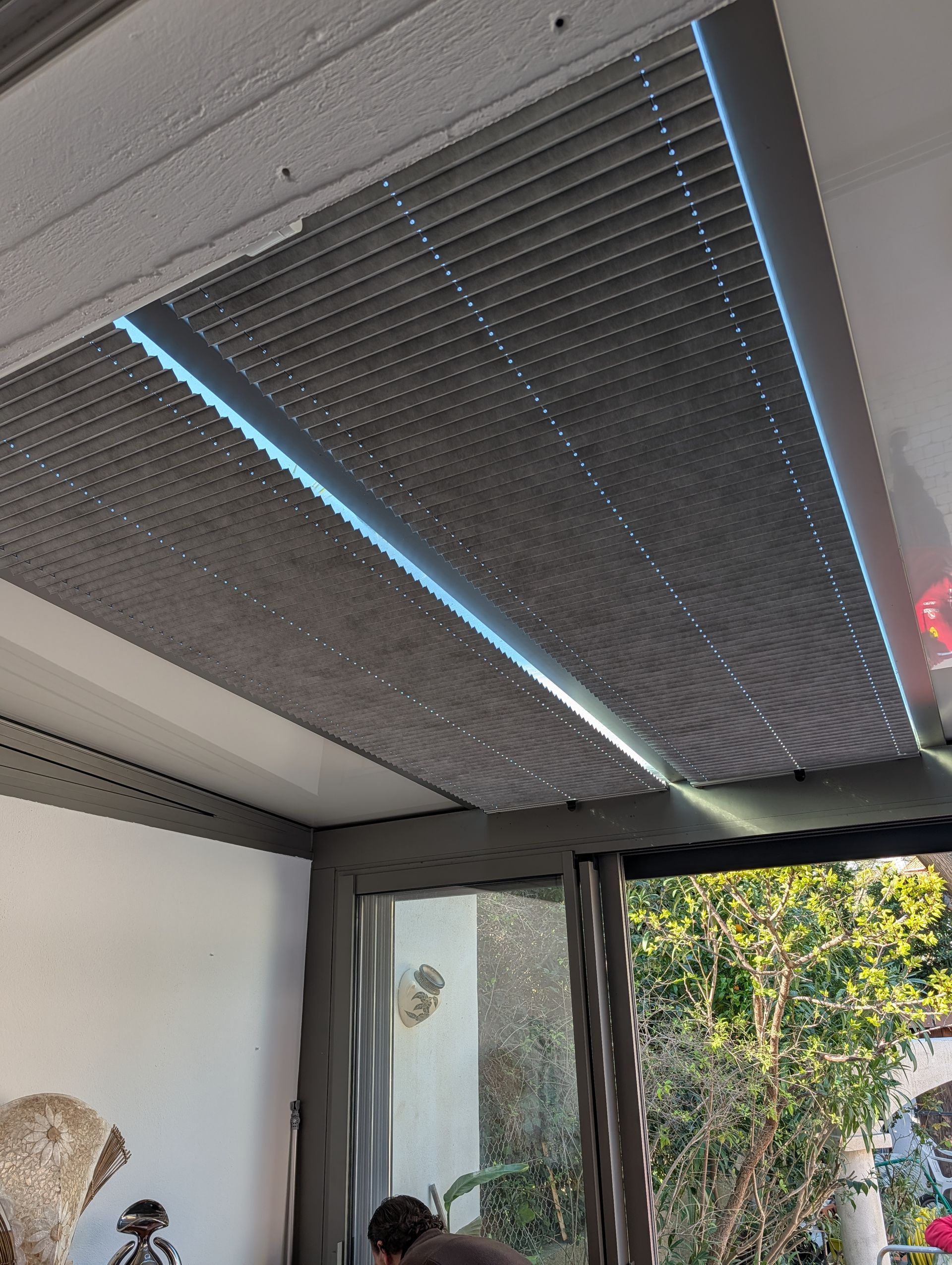 Toit de pergola extérieur rétractable gris foncé laissant filtrer la lumière à travers les lattes, vu de l'intérieur d'une maison.