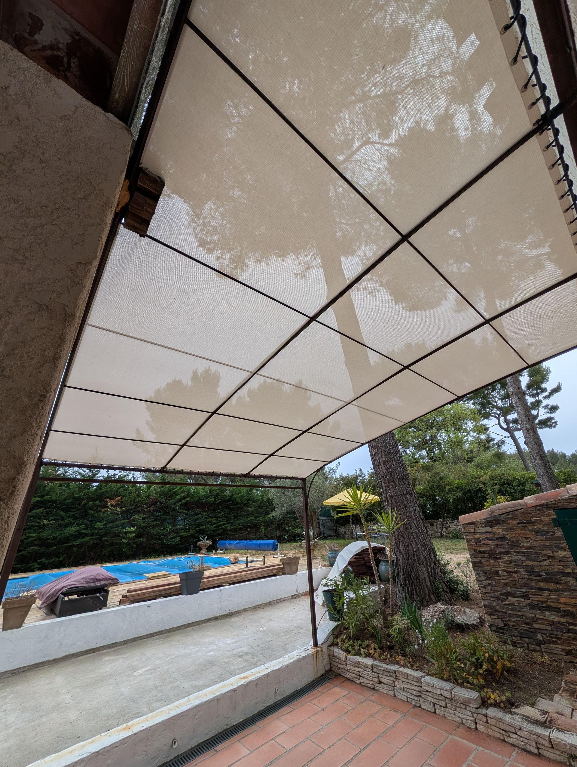 Une pergola beige à motif quadrillé s'étend au-dessus d'une terrasse donnant sur un jardin avec une piscine visible en arrière-plan.