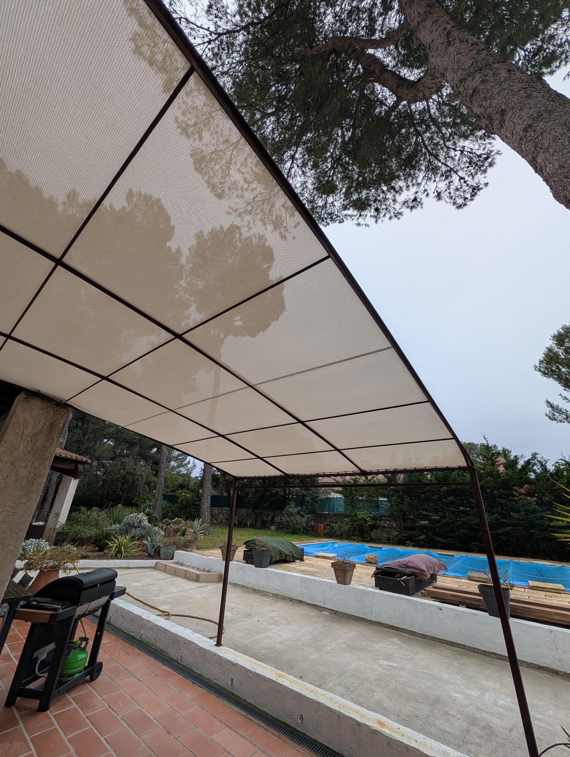 Une voile d'ombrage rectangulaire beige fixée à un bâtiment, s'étendant au-dessus d'une terrasse avec une piscine et des arbres à l'arrière.