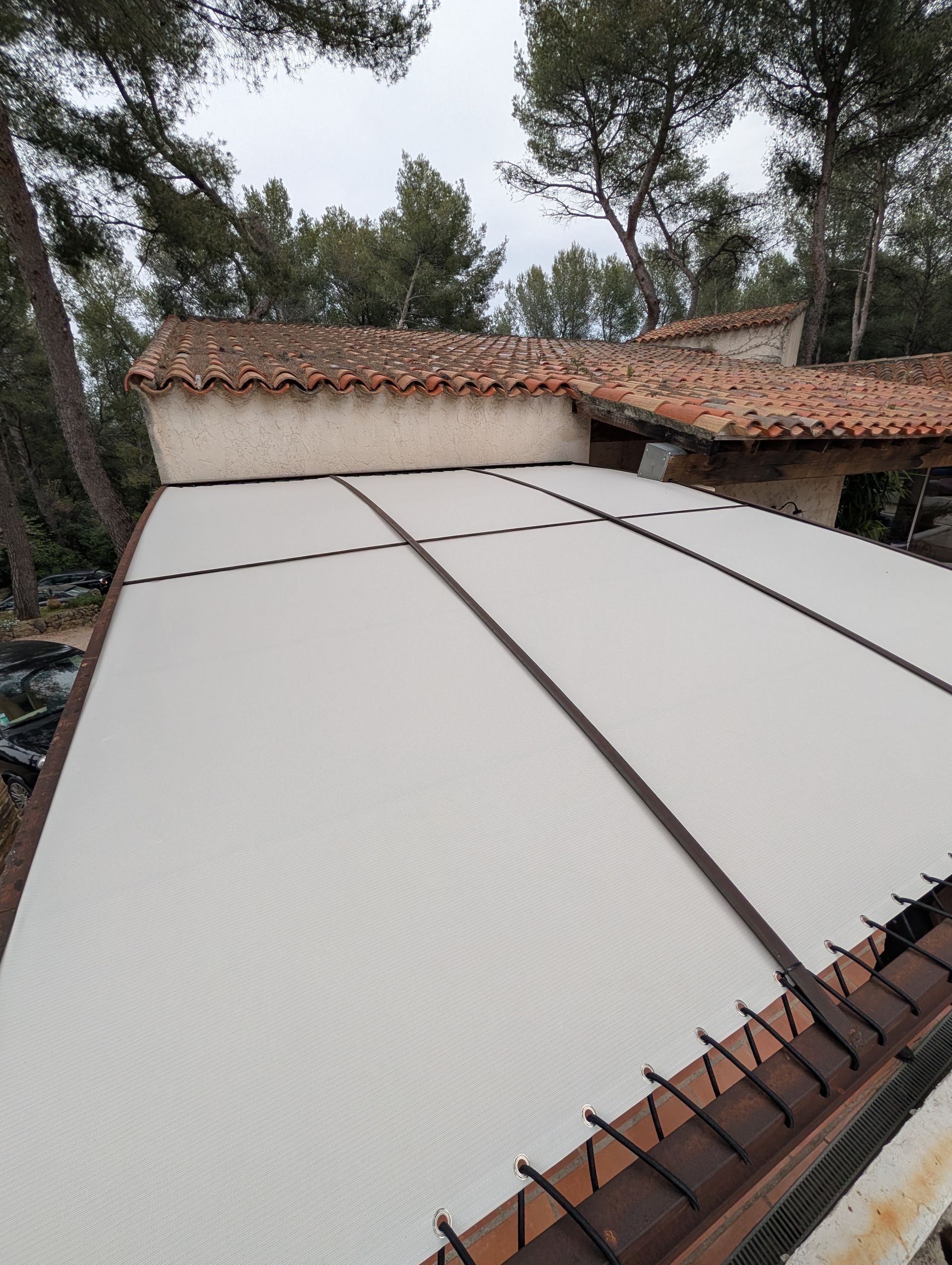 Un auvent rétractable couleur crème recouvre un patio, adossé à un bâtiment rustique en pierre au toit de tuiles en terre cuite.
