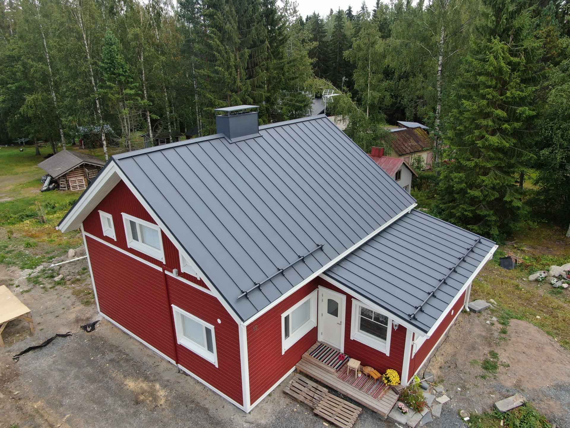 Punainen talo harmaalla metallikatolla, mäntyjen ympäröimänä maaseudun aukiolla