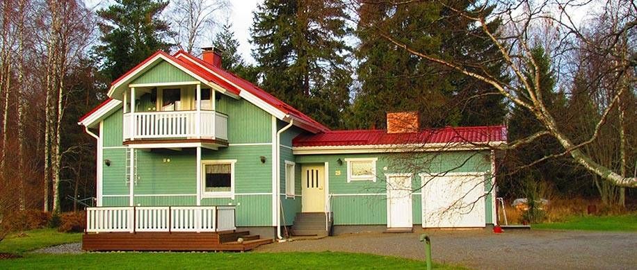 Pastellinvihreä talo punaisella katolla ja kuistilla ajotien vieressä metsäisellä pihalla
