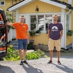 Kaksi miestä seisoo keltaisen talon ulkopuolella mustan kuorma-auton vieressä, toinen oranssissa ja toinen violetissa paidassa