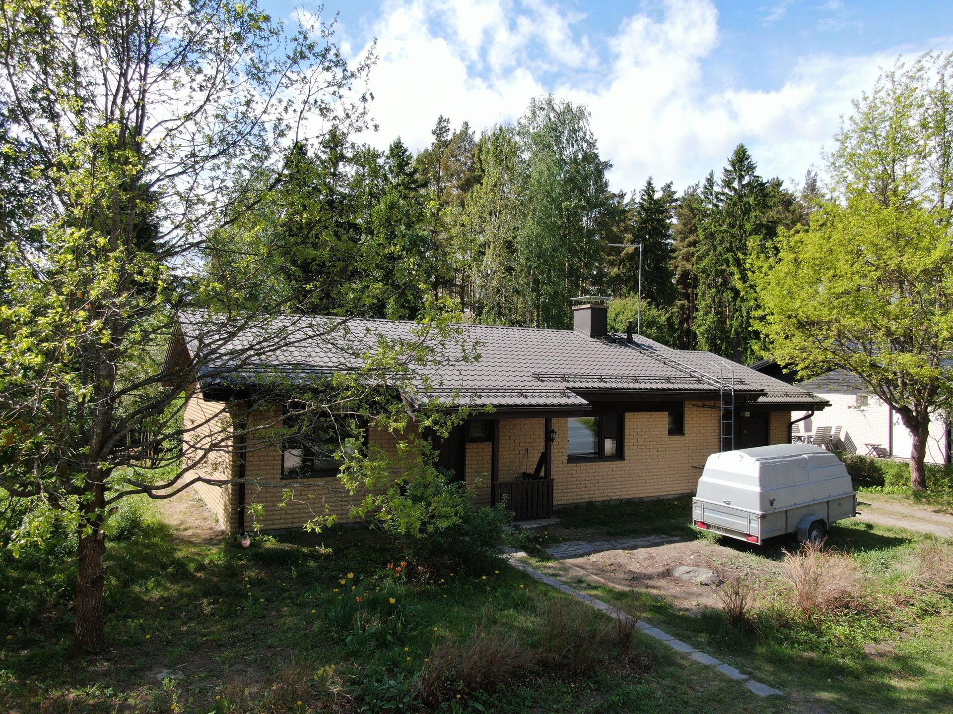 Pieni harmaakattoinen tiilitalo metsäisellä pihalla, jonka ulkopuolella on pysäköity valkoinen pakettiauto.