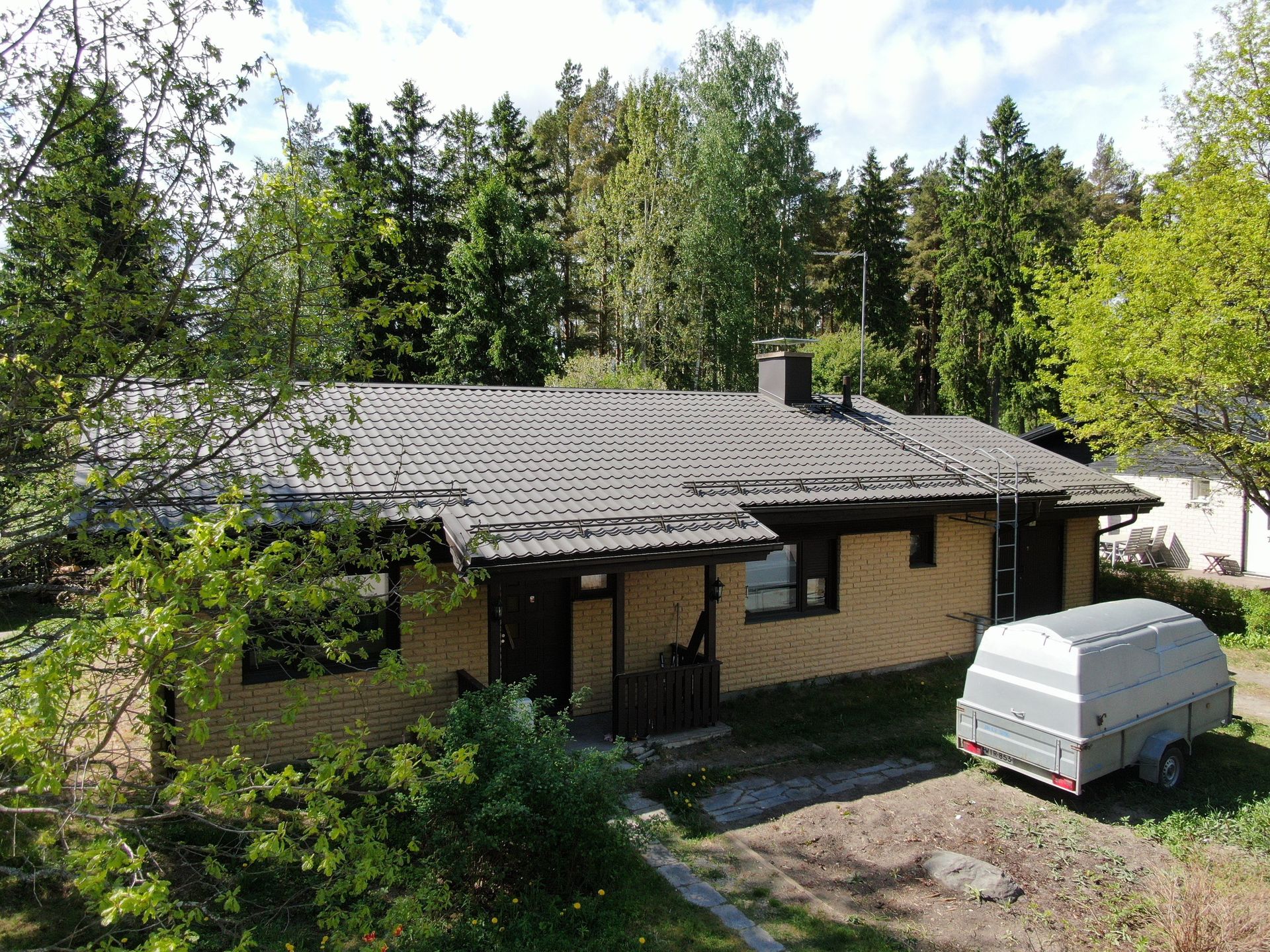 Pieni tummakattoinen talo metsäisellä pihalla, jonka ulkopuolella on pysäköity valkoinen pakettiauto.