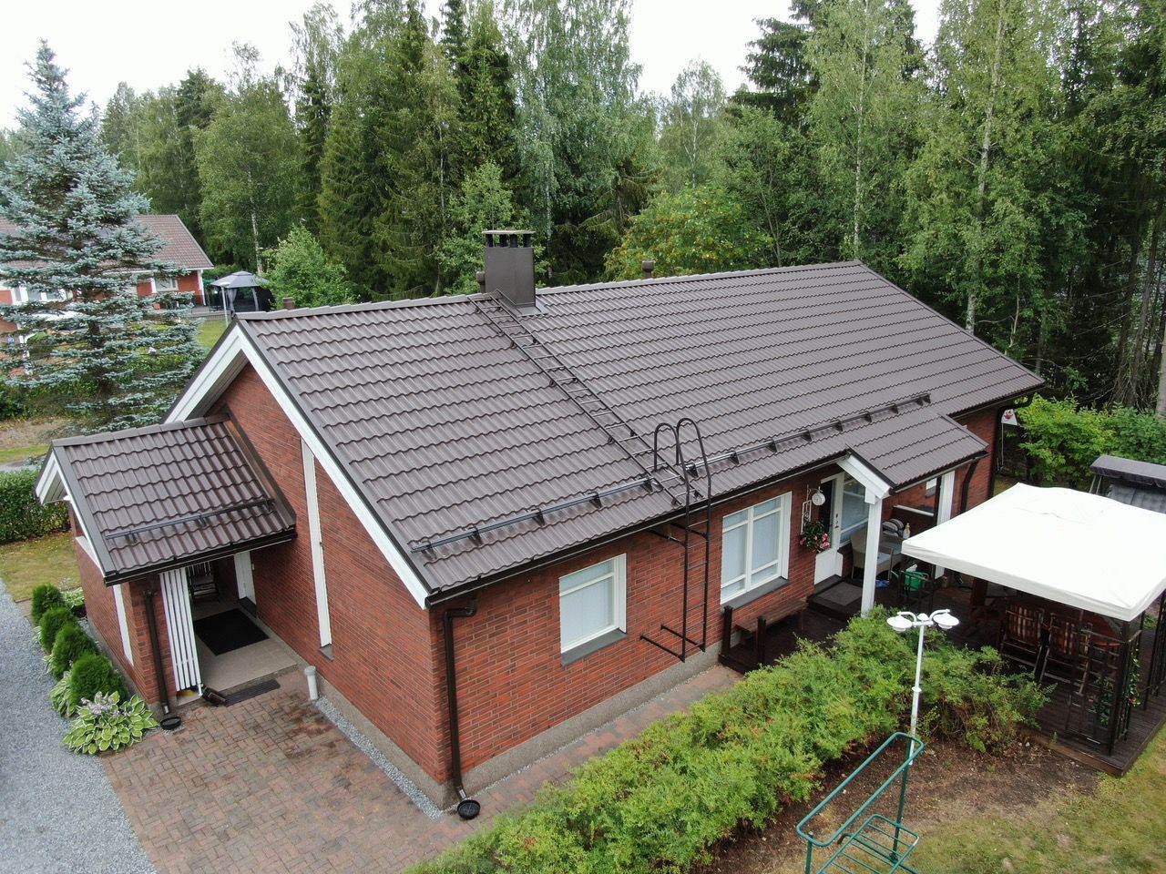 Ilmakuva punatiilisestä talosta, jossa on ruskea katto, puiden ja pienen puutarhan ympäröimänä.