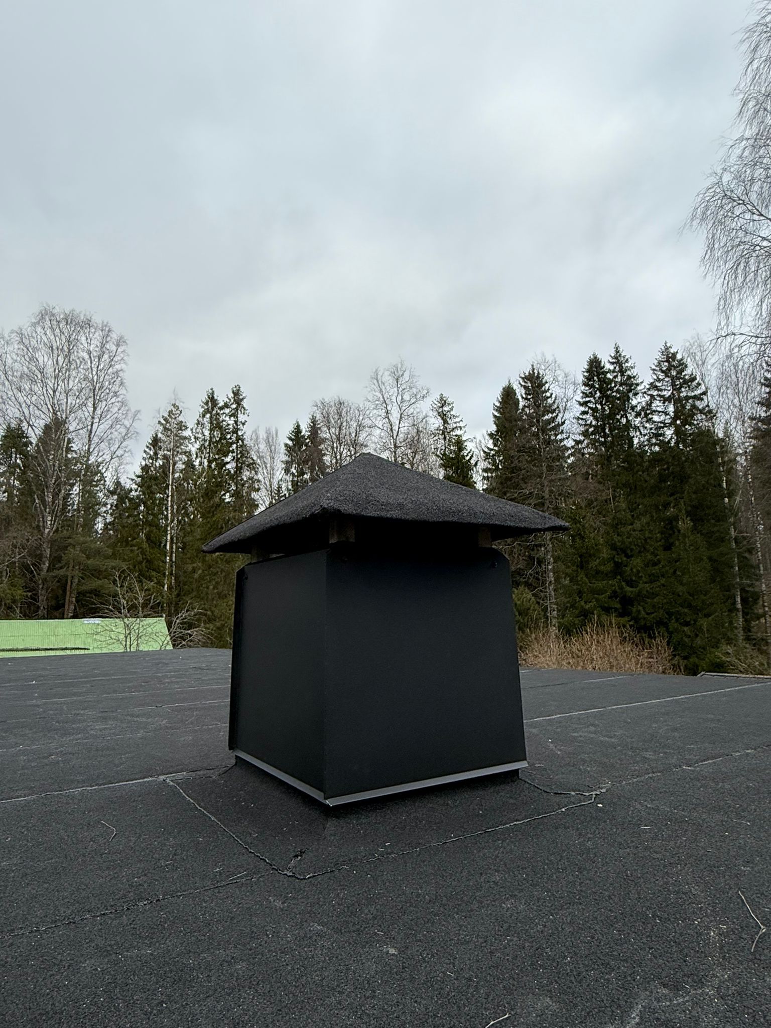 Musta kattotuuletusaukko sorapinnalla, taustalla puita ja pilvinen taivas