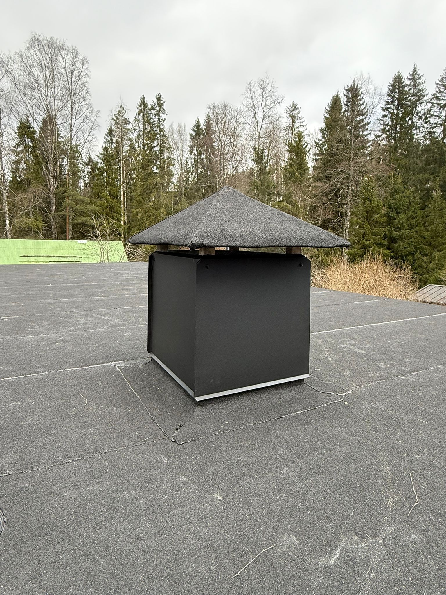 Musta ulkolaatikko, jossa on kalteva katto soralla, puita ja pilvistä taivasta vasten