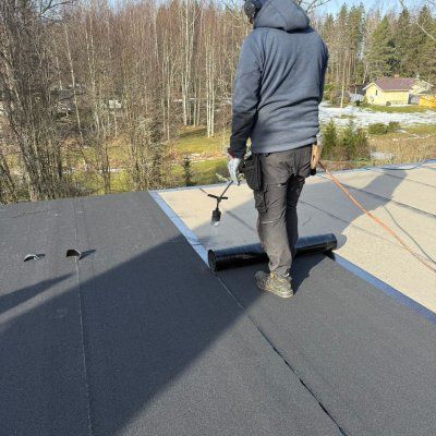 Työntekijä rullaa mustaa kattokalvoa kaltevalla katolla ulkona
