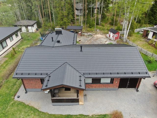 Ilmakuva tummanharmaakattoisesta tiilitalosta, jossa on katettu kuisti ja sorapiha.
