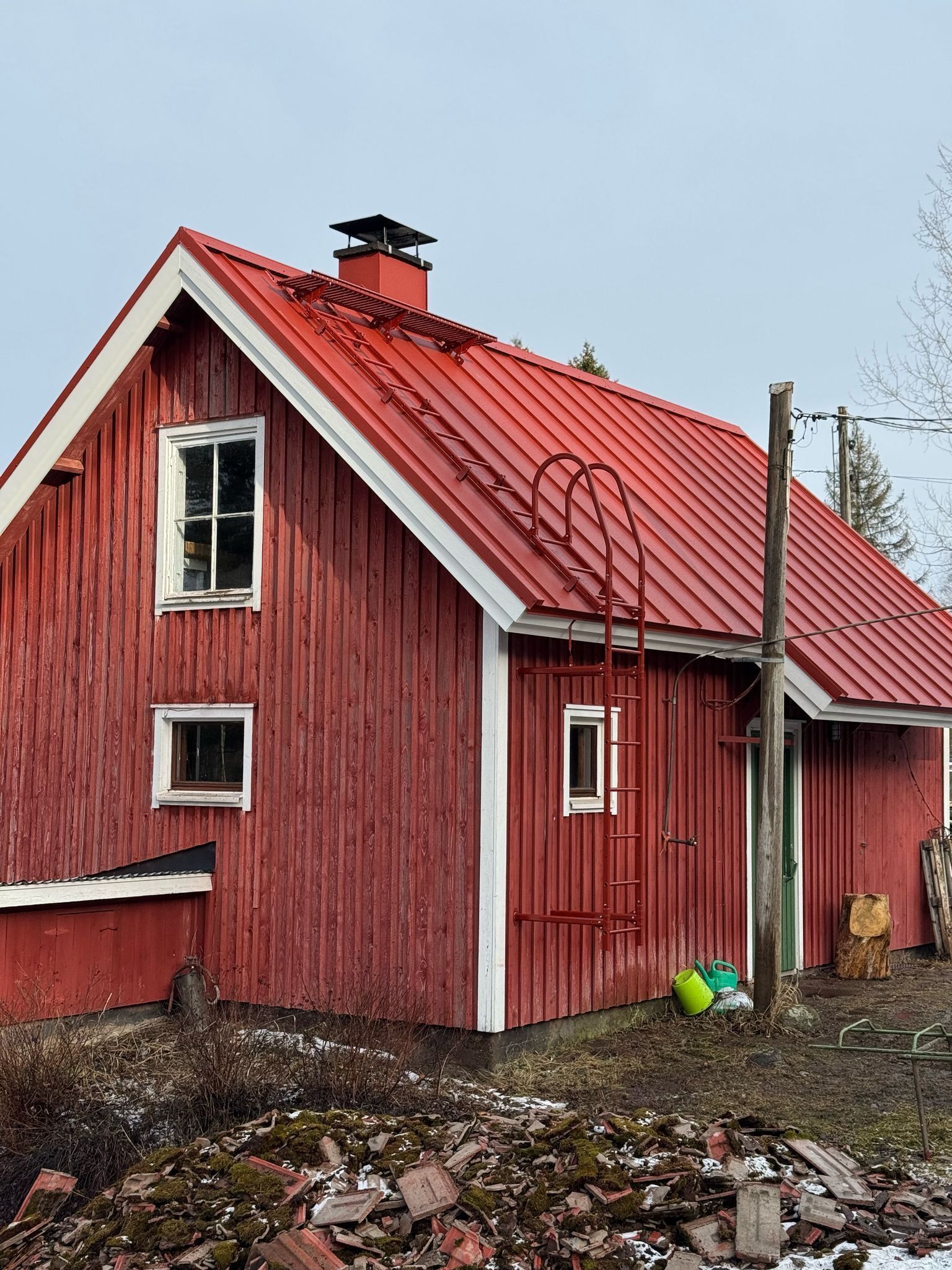 Punainen puutalo valkoisilla reunuksilla ja punaisella metallikatolla mutaisella tontilla harmaan taivaan alla