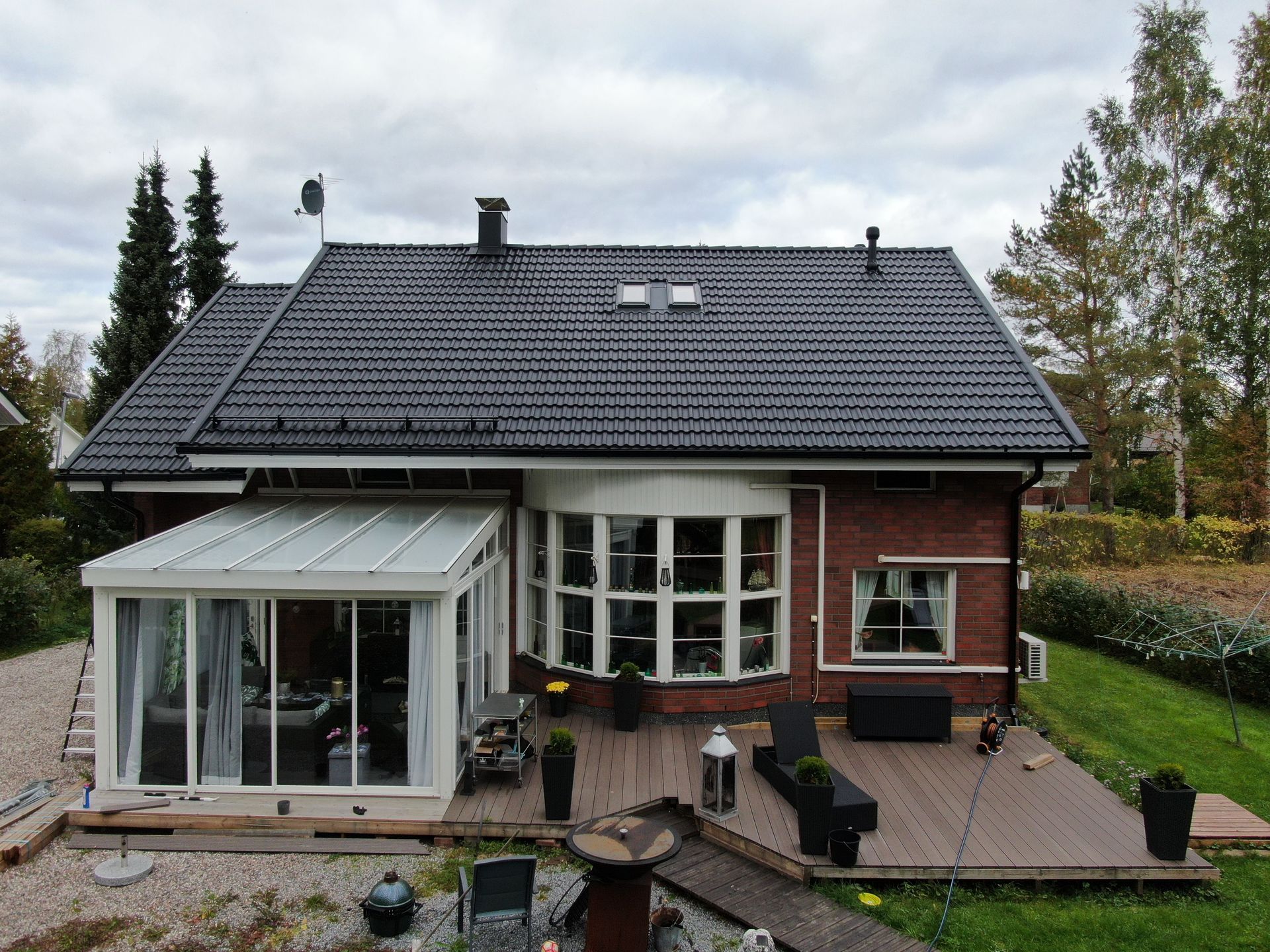 Erillinen tiilitalo, jossa on tumma katto, lasinen aurinkohuone ja kivetty etuterassi esikaupunkipuutarhassa