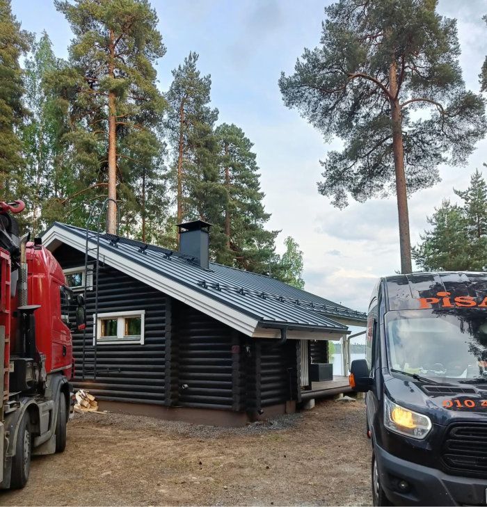 Pieni musta mökki mäntymetsässä, jonka molemmille puolille on pysäköity kaksi kuorma-autoa