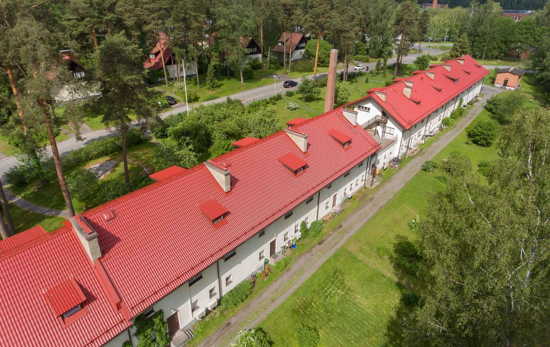 Ilmakuva pitkistä valkoisista, punakattoisista rakennuksista puiden ja tien vieressä.