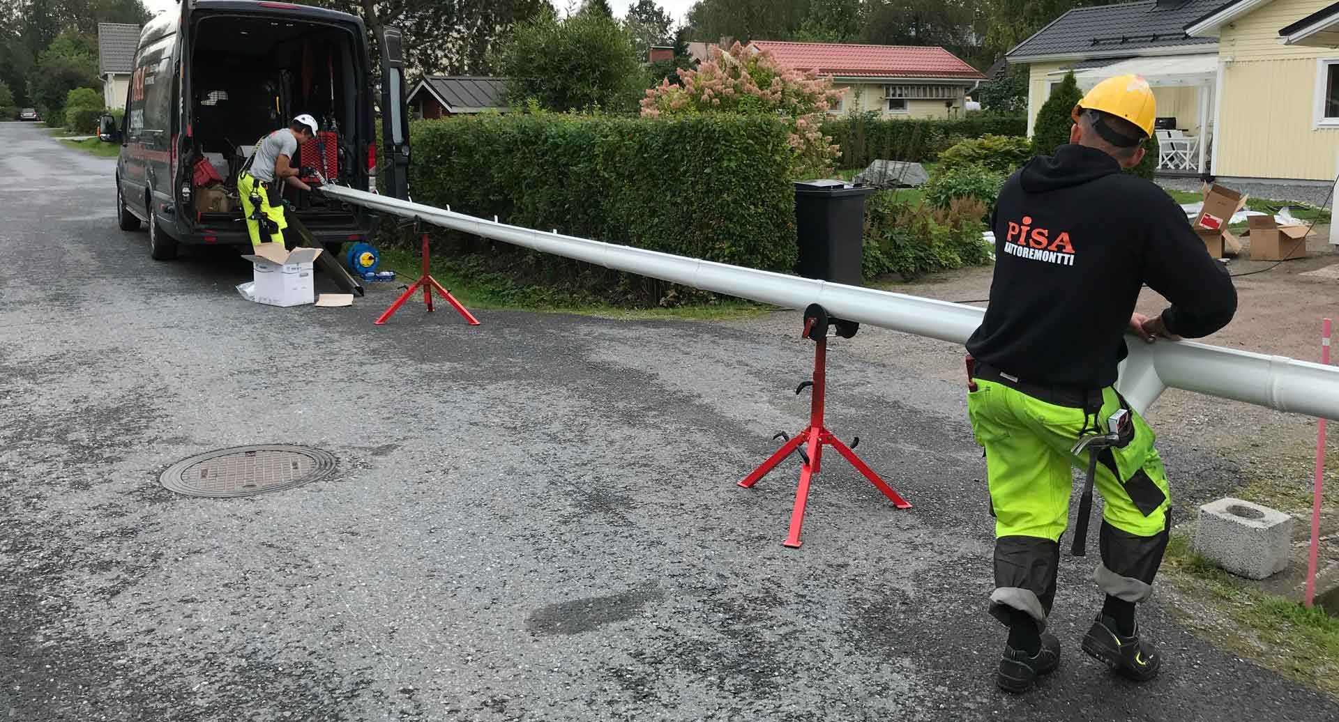 Työntekijät asentavat pitkää metalliputkea märän kadun poikki kuorma-auton ja punaisten putkitukien viereen