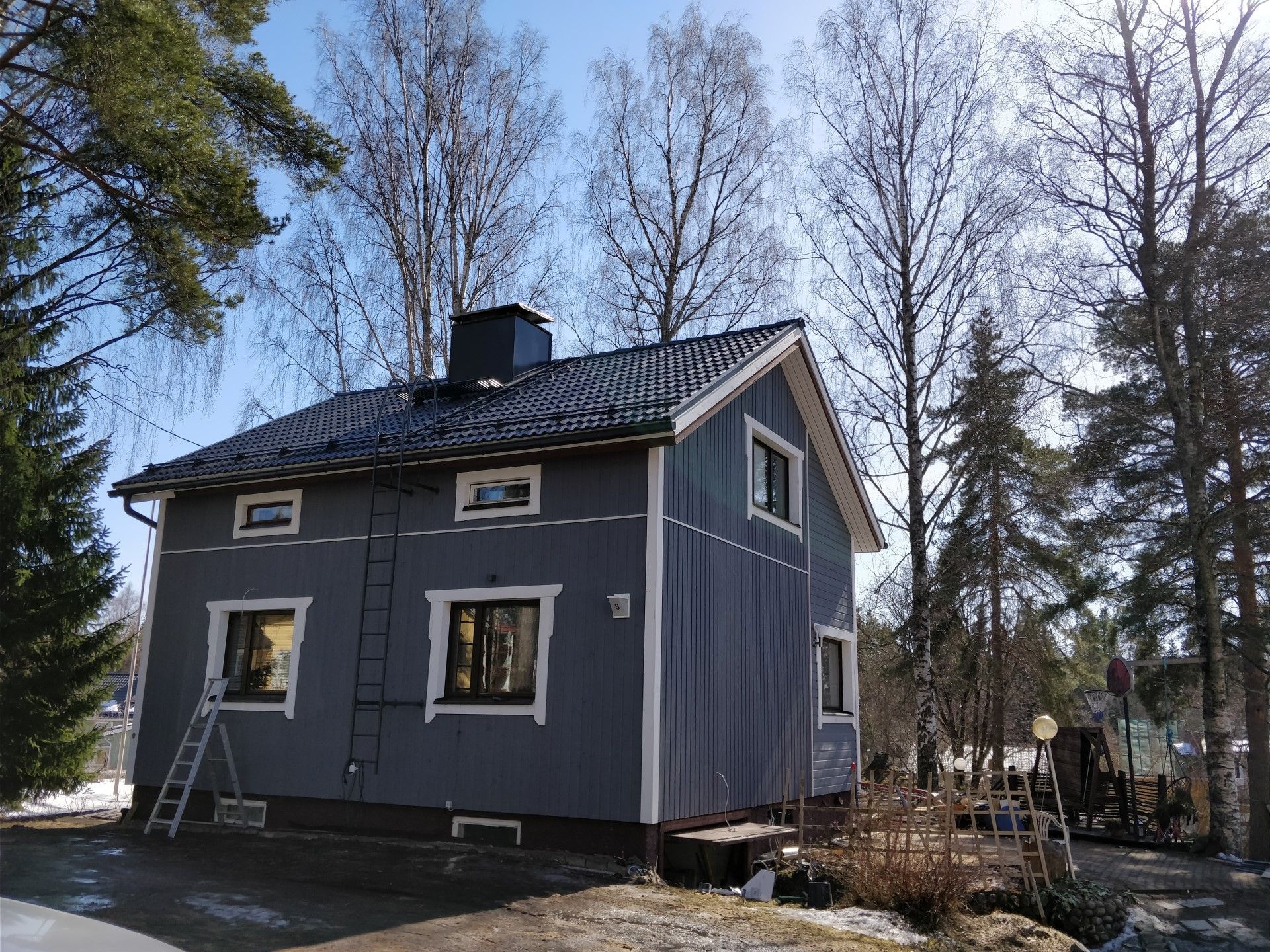 Harmaa kaksikerroksinen talo valkoisilla reunuksilla ja metallikatolla lehdettömien puiden keskellä metsäisellä pihalla