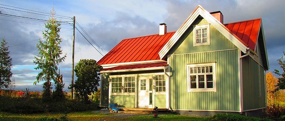 Vihreä talo punaisella katolla aurinkoisella maaseudun pihalla, puiden ja pilvisen taivaan kanssa.