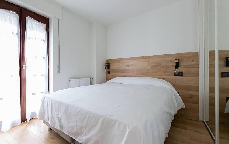 Dormitorio con cama blanca, cabecero de madera y armario con espejo. Ventana con cortinas a la izquierda.