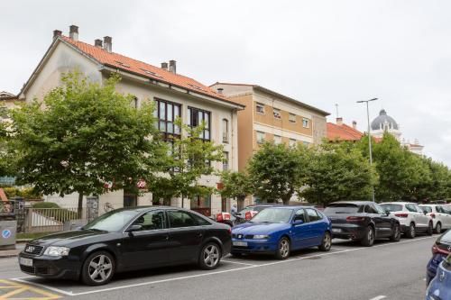 Coches aparcados en una calle frente a un edificio de color claro con árboles y una cúpula a lo lejos.