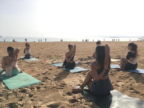 Personas practicando yoga en la playa; día soleado, colchonetas, estiramientos de brazos, océano al fondo.