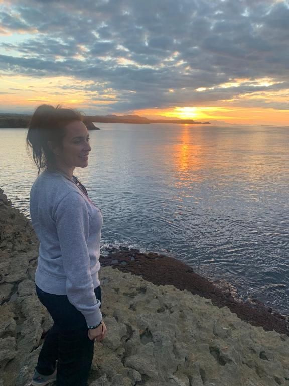 Una mujer de pie en un acantilado contempla la puesta de sol sobre el océano. El cielo anaranjado y amarillo se refleja en el agua.