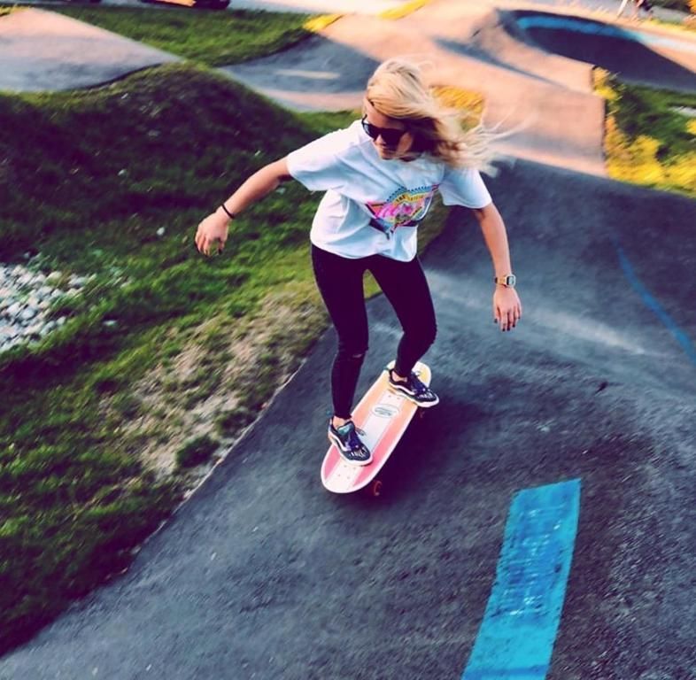 Mujer en monopatín bajando por una pista pavimentada con colinas de hierba.