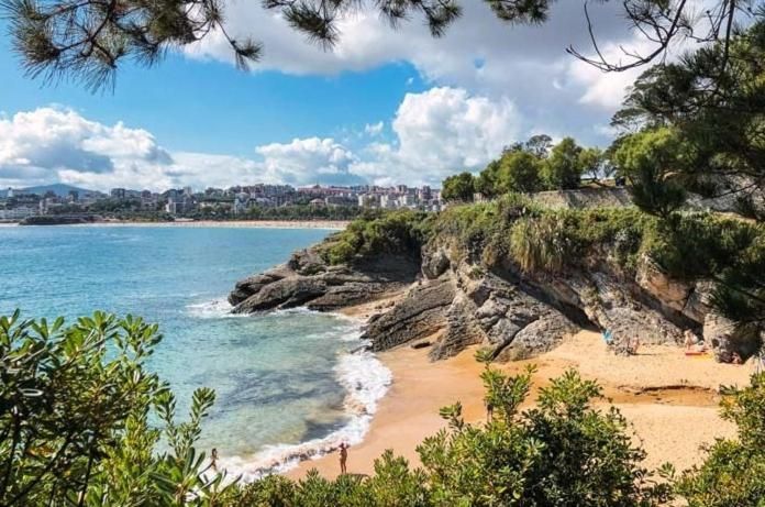 Escena de playa: orilla arenosa, acantilados rocosos, océano azul, ciudad costera al fondo, cielo azul brillante.