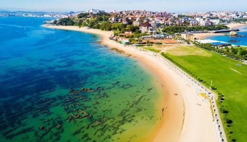 Vista aérea de una playa de arena que se curva a lo largo de aguas turquesas, con una zona verde y edificios de la ciudad al fondo.